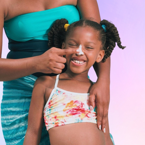 African American girl in a swimsuit with white sunscreen being applied to her nose by a woman in a blue dress